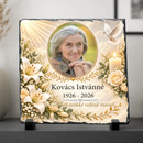 Négyzet alakú, rusztikus szélű palakő kegyeleti fotókő fekete talpakon. Közepén egy idős hölgy ovális fotója látható arany szegéllyel, mellette fehér liliomok, krémszínű rózsák, égő gyertya, fehér galamb és égi fénysugarak.
