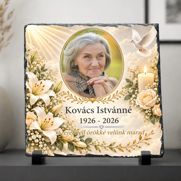 Négyzet alakú, rusztikus szélű palakő kegyeleti fotókő fekete talpakon. Közepén egy idős hölgy ovális fotója látható arany szegéllyel, mellette fehér liliomok, krémszínű rózsák, égő gyertya, fehér galamb és égi fénysugarak.